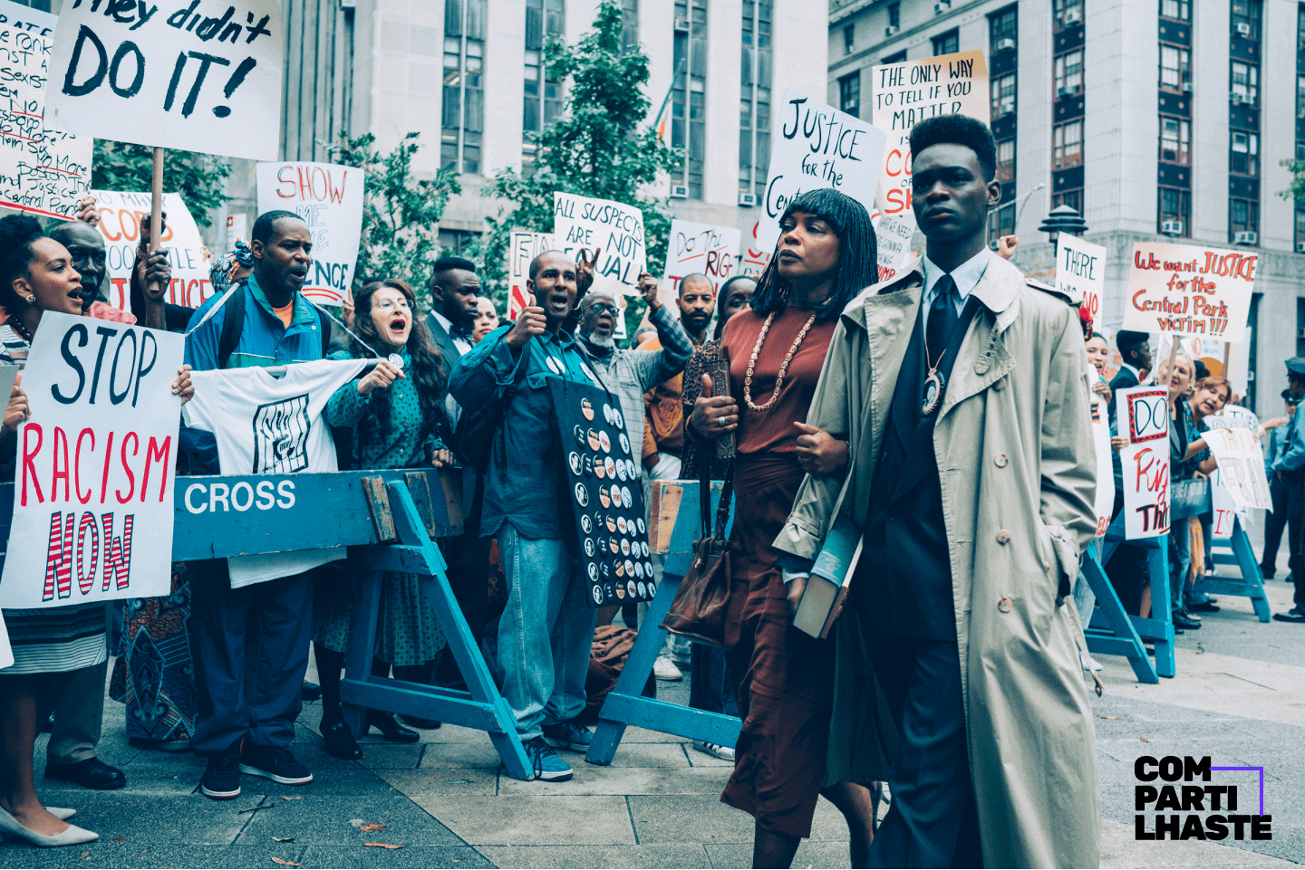 Foto de uma cena da série Olhos que condenam, em que um jovem negro está andando ao lado da mãe enquanto acontece um protesto contra o racismo ao lado.